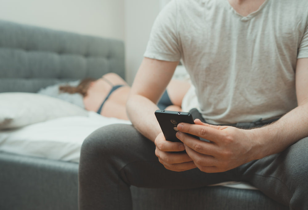 Man Texting Sitting On The Edge Of A Bed While His Partner Sleeps